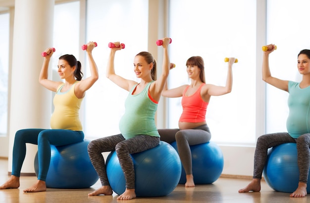 embarazo, deporte, fitness, personas y concepto de estilo de vida saludable - grupo de mujeres embarazadas felices con pesas haciendo ejercicio en la pelota en el gimnasio