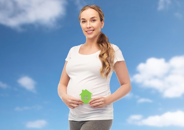 el embarazo, la ecología, la gente y el concepto de vivienda - mujer embarazada feliz con una casa verde sobre el cielo azul y el fondo de las nubes