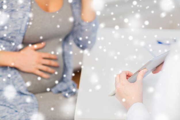 Embarazo, invierno, medicina, salud y concepto de personas: cierre de un médico ginecólogo que muestra una computadora de tablet pc a una mujer embarazada en el hospital sobre la nieve