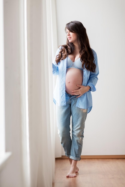 Embarazo, maternidad, personas y concepto de expectativas: mujer embarazada feliz con una gran barriga desnuda mirando por la ventana en casa