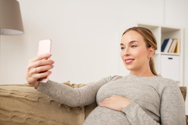 embarazo, maternidad, tecnología, personas y concepto de expectativas - mujer embarazada feliz con teléfono inteligente tomando selfies en casa