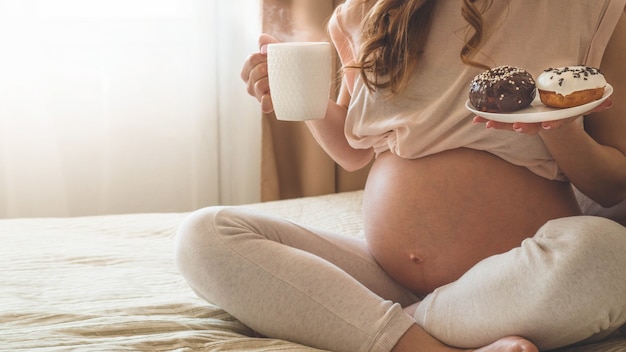 Embarazo y nutrición. Mujer embarazada disfrutando de donas y té en la cama, espacio libre