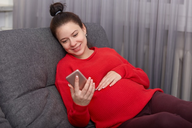 Embarazo, ocio, concepto de tecnología. Feliz sonriente niña embarazada tiene teléfono inteligente, usa suéter rojo, descansa en el sofá