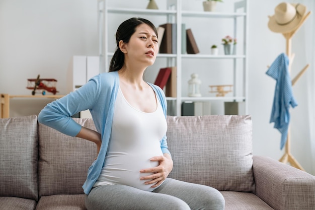 embarazo personas de salud y concepto de expectativa. joven asiática embarazada en el sofá tocándose la espalda y sufriendo de dolor de espalda en casa. madre con el ceño fruncido con el bebé en el vientre sentado en el sofá.