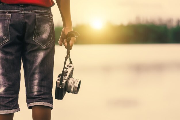 Embrome al fotógrafo del muchacho que sostiene una cámara del vintage en el parque de la orilla.