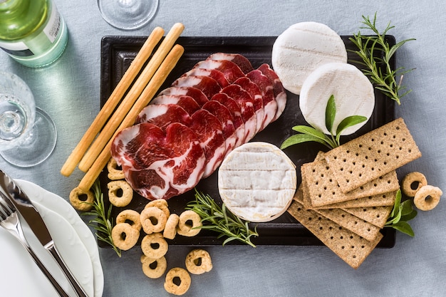 Los embutidos y el queso se sirven en una bandeja sobre una mesa con vino blanco, galletas, grissini y taralli con hierbas aromáticas sobre un mantel festivo de lino azul.