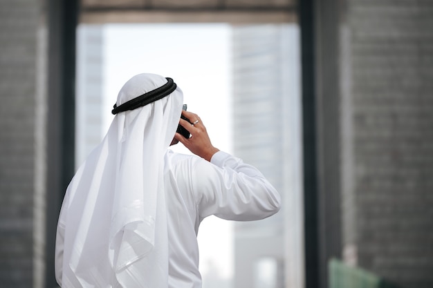 Emirati vestindo kandura tradicional falando com telefone inteligente.