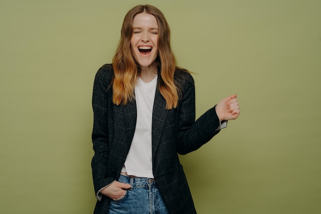 Emoción. Disparo de estudio de mujer joven sorprendida y feliz con cabello castaño ondulado posando en chaqueta formal oscura, top blanco y jeans sobre fondo verde con una mano en el bolsillo. Logros personales