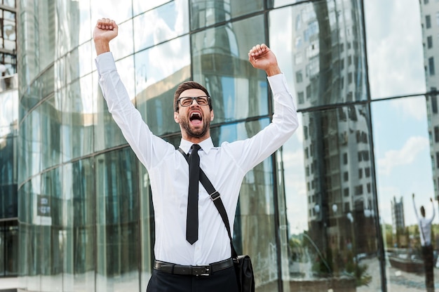 Foto emoción empresarial. alegre joven empresario manteniendo los brazos en alto y expresando positividad mientras está de pie al aire libre
