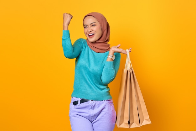Emocionada y alegre joven asiática sosteniendo bolsas de compras sobre fondo amarillo