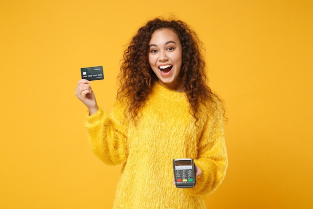 Emocionada chica afroamericana posando aislada sobre fondo amarillo. Concepto de estilo de vida de las personas. Simulacros de espacio de copia. Sosteniendo una terminal de pago bancaria inalámbrica moderna para procesar la adquisición de pagos con tarjeta de crédito.