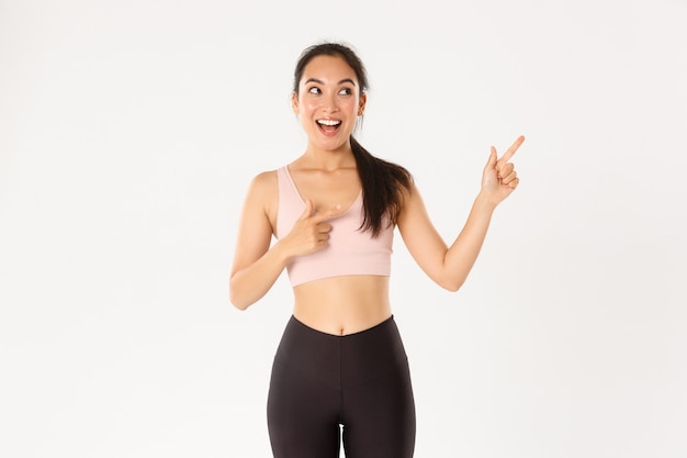 Emocionada chica de fitness asiática delgada y linda, atleta femenina apuntando con el dedo en la esquina superior derecha.