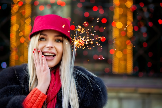 Emocionada chica rubia sonriente con traje de invierno de moda, divirtiéndose con luces de bengala en el fondo bokeh. Espacio para texto