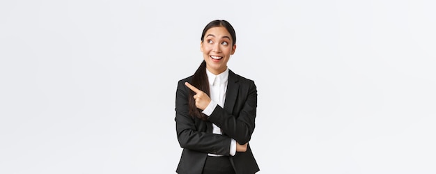 Emocionada y divertida mujer de negocios asiática joven en traje apuntando con el dedo a la izquierda y mirando emocionada por el anuncio promocional de fondo blanco Empresario haciendo una gran sugerencia