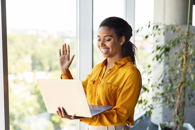 Emocionada empresaria afroamericana que tiene una videollamada en una computadora portátil saludando con la mano a la cámara web de pie cerca de la ventana