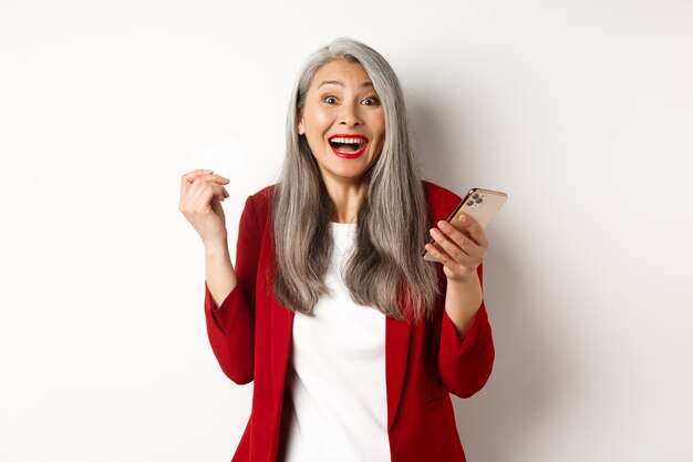 Emocionada empresaria asiática en chaqueta roja, sosteniendo una tarjeta de crédito de plástico y un teléfono inteligente, sonriendo feliz a la cámara, de pie sobre fondo blanco.