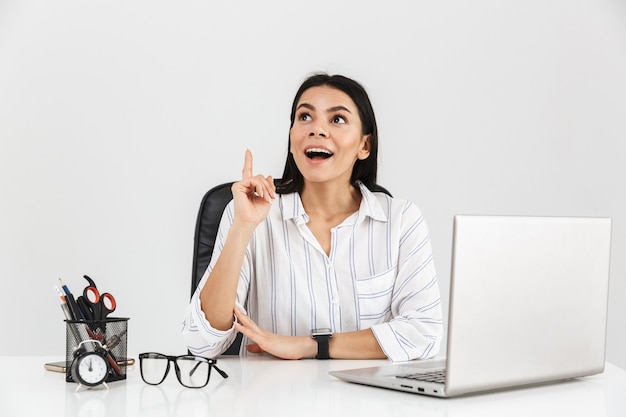 Emocionada empresaria morena sentada a la mesa con papelería y trabajando en la computadora portátil en la oficina aislada sobre la pared blanca