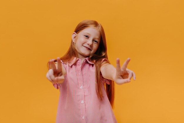 Emocionada y encantadora chica pelirroja con camisa a rayas sonriente El niño muestra el signo de la paz en un fondo naranja aislado