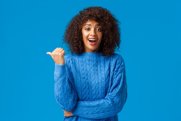 Emocionada feliz bonita mujer afroamericana con corte de pelo afro en suéter de invierno, apuntando con el pulgar a la izquierda y riendo mientras discute el momento divertido, hablando casualmente durante la fiesta, pared azul