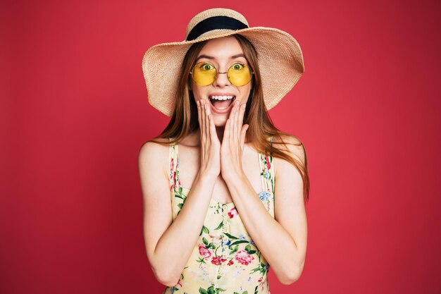 Emocionada y feliz, hermosa y encantadora mujer joven con gafas de sol, vestido y sombrero de verano se divierte mientras posa en un fondo rosa