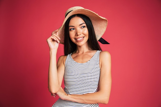 Emocionada y feliz, hermosa y encantadora mujer joven con gafas de sol, vestido y sombrero de verano se divierte mientras posa en un fondo rosa