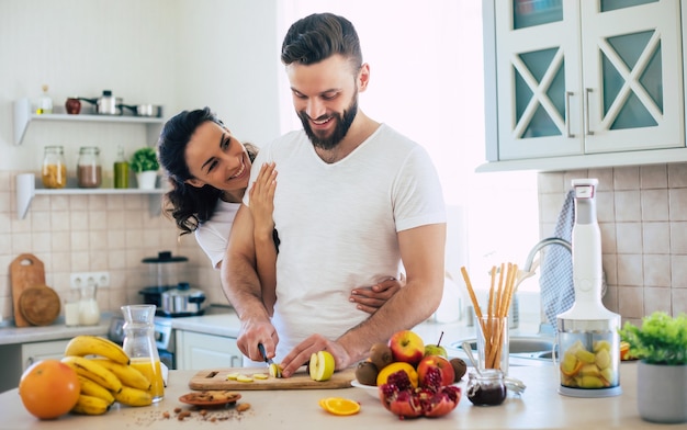 Emocionada feliz hermosa joven pareja enamorada de cocinar en la cocina y divertirse juntos mientras hace ensalada de frutas frescas y saludables