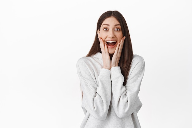 Emocionada y feliz mujer joven no puede creer que ganó, tocando la cara y sonriendo, luciendo asombrada y emocionada con cara de sorpresa, ganando, de pie sobre una pared blanca