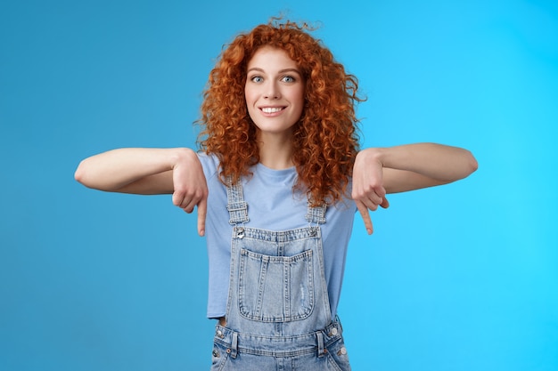 Emocionada, feliz, sonriente, cariñosa, pelirroja, novia de pelo rizado, emocionada esperando viaje de vacaciones de verano, apuntando hacia abajo, dedo índice, estado de ánimo optimista, mostrar tienda favorita, comprar mejores precios, fondo azul.