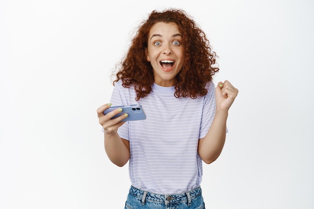 Foto emocionada ganadora, mujer pelirroja sosteniendo un teléfono móvil y luciendo asombrada como si ganara, se convierte en campeona en videojuegos, fondo blanco. copia espacio