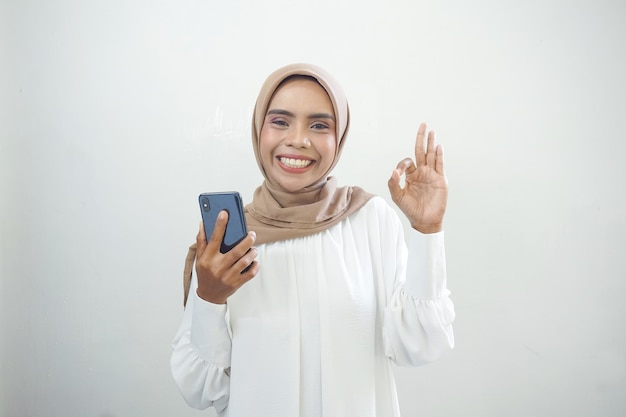 Emocionada hermosa mujer musulmana asiática mostrando teléfono móvil aislado sobre fondo blanco.