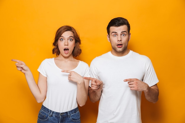 Emocionada hermosa pareja vistiendo camisetas blancas que se encuentran aisladas sobre la pared amarilla, apuntando al espacio de la copia