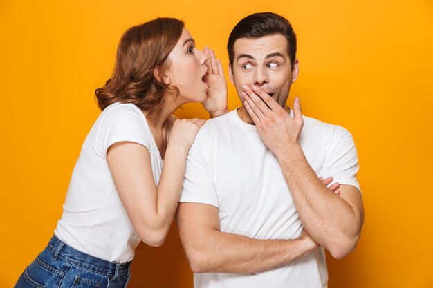 Foto emocionada hermosa pareja vistiendo camisetas blancas que se encuentran aisladas sobre la pared amarilla, contando secretos