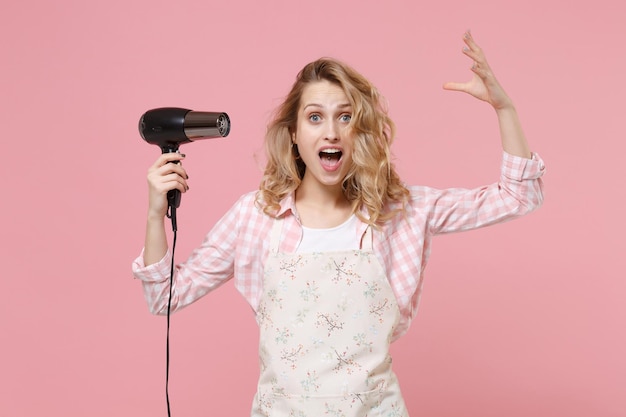 Emocionada joven ama de casa con ropa informal, delantal posando aislada en un fondo rosa pastel en el estudio. Concepto de limpieza. Simulacros de espacio de copia. Secar el cabello con secador de pelo, extendiendo las manos.