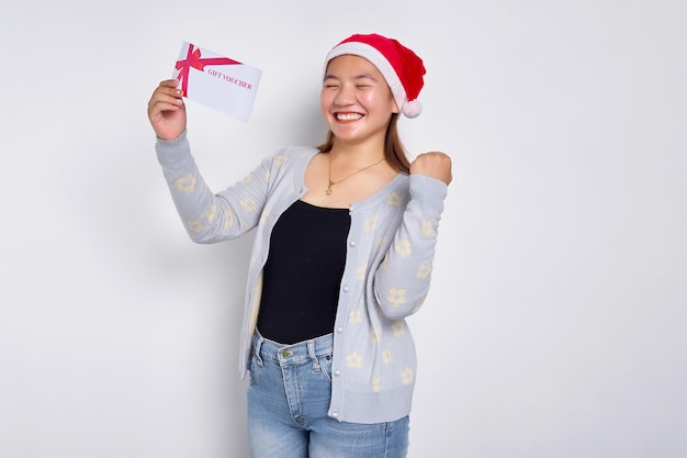 Emocionada joven asiática de 20 años con sombrero de Navidad sosteniendo una tarjeta de cupón de cupón de regalo para la tienda haciendo un gesto de ganador mirando una cámara aislada en el fondo blanco del estudio