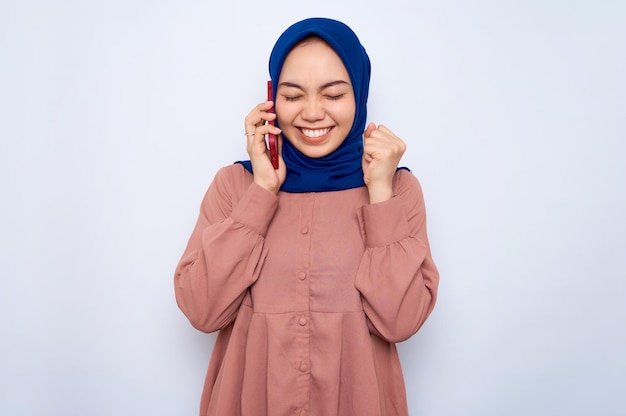 Emocionada joven asiática con camisa rosa hablando por teléfono móvil y haciendo un gesto de ganador aislado sobre fondo blanco Concepto de estilo de vida religioso de la gente