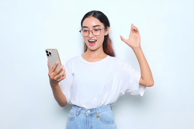 Emocionada joven asiática con camiseta blanca usando teléfono móvil celebrando el éxito recibiendo buenas noticias aisladas de fondo blanco