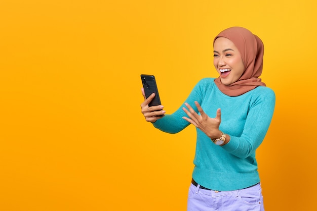 Emocionada joven asiática con smartphone celebrando el éxito aislado sobre fondo amarillo