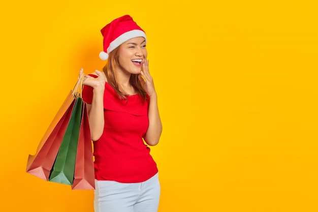 Emocionada joven asiática con sombrero de Santa Claus sosteniendo bolsas de la compra y mirando a otro lado en el espacio de la copia