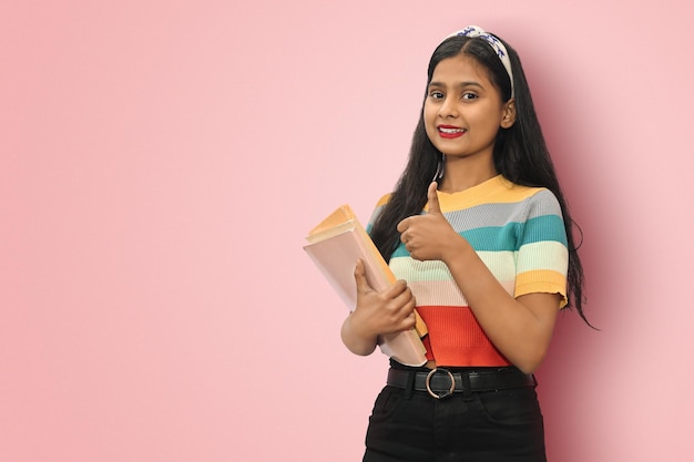 Emocionada joven estudiante india asiática de pie de lado posando aislada sosteniendo libros de texto y mostrando el pulgar hacia arriba mirando directamente a la cámara mujer de cabello oscuro expresando emociones positivas