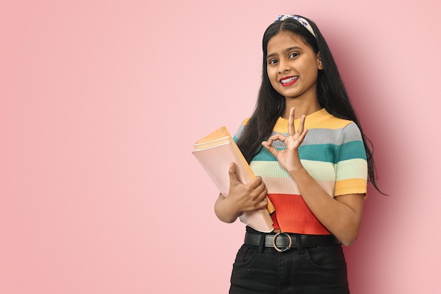 Emocionada joven estudiante india asiática de pie de lado posando aislada sosteniendo libros de texto y mostrando un signo de estar bien mirando directamente a la cámara mujer de cabello oscuro expresando emociones positivas