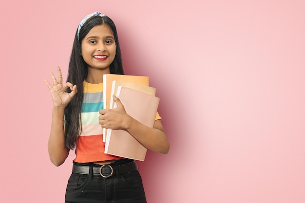 Emocionada joven estudiante india asiática posando aislada sosteniendo libros de texto y mostrando el signo de ok mirando directamente a la cámara mujer de cabello oscuro expresando emociones positivas