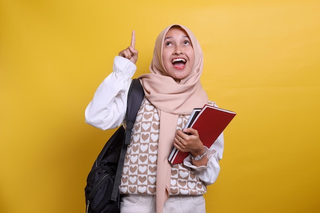 Emocionada joven estudiante musulmana asiática en hijab mostrando una expresión feliz con el dedo apuntando hacia arriba wh