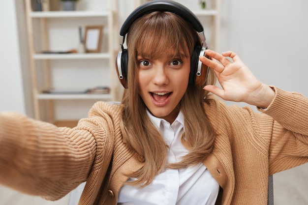 Emocionada joven estudiante rubia independiente con suéter cálido en los auriculares escucha canciones favoritas sentada en un sillón en el interior de una casa moderna Tiempo de música Relajante Lista de reproducción fresca Concepto Amplia vista de selfie