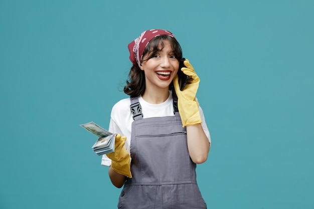 Emocionada joven limpiadora con pañuelo uniforme y guantes de goma sosteniendo dinero mirando la cámara mientras habla por teléfono aislada en el fondo azul