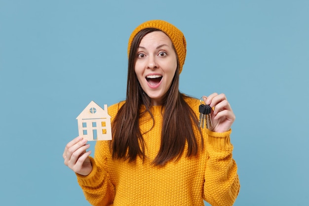 Foto emocionada joven morena con suéter amarillo y sombrero posando aislada en un retrato de estudio de fondo azul. gente emociones sinceras concepto de estilo de vida. simulacros de espacio de copia. mantenga el manojo de llaves de la casa.