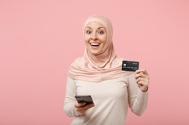 Emocionada joven mujer musulmana árabe con ropa ligera hiyab posando aislada en un fondo rosa. Concepto de estilo de vida del Islam religioso de la gente. Simulacros de espacio de copia. Usando el teléfono móvil, sostenga la tarjeta bancaria de crédito.