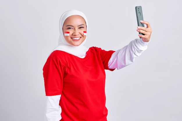 Emocionada joven musulmana asiática con camiseta blanca roja sosteniendo un teléfono móvil y celebrando el día de la independencia de Indonesia el 17 de agosto aislada de fondo blanco