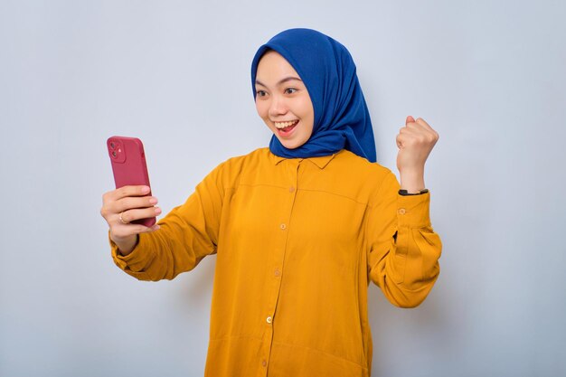 Emocionada joven musulmana asiática vestida con pantalones naranja usando un teléfono móvil celebrando el éxito recibiendo buenas noticias aisladas sobre fondo blanco