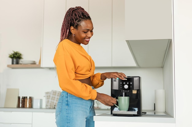 Emocionada joven negra haciendo café aromático fresco en una máquina moderna en el interior de la cocina