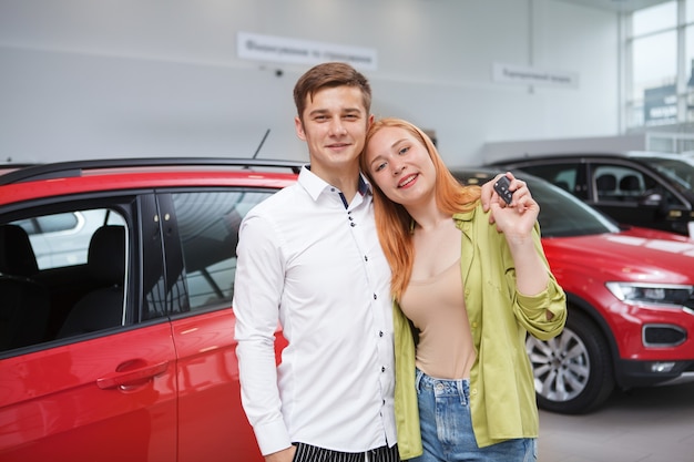 Emocionada joven pareja abrazándose, mostrando la llave del coche a la cámara después de comprar un automóvil nuevo en el concesionario